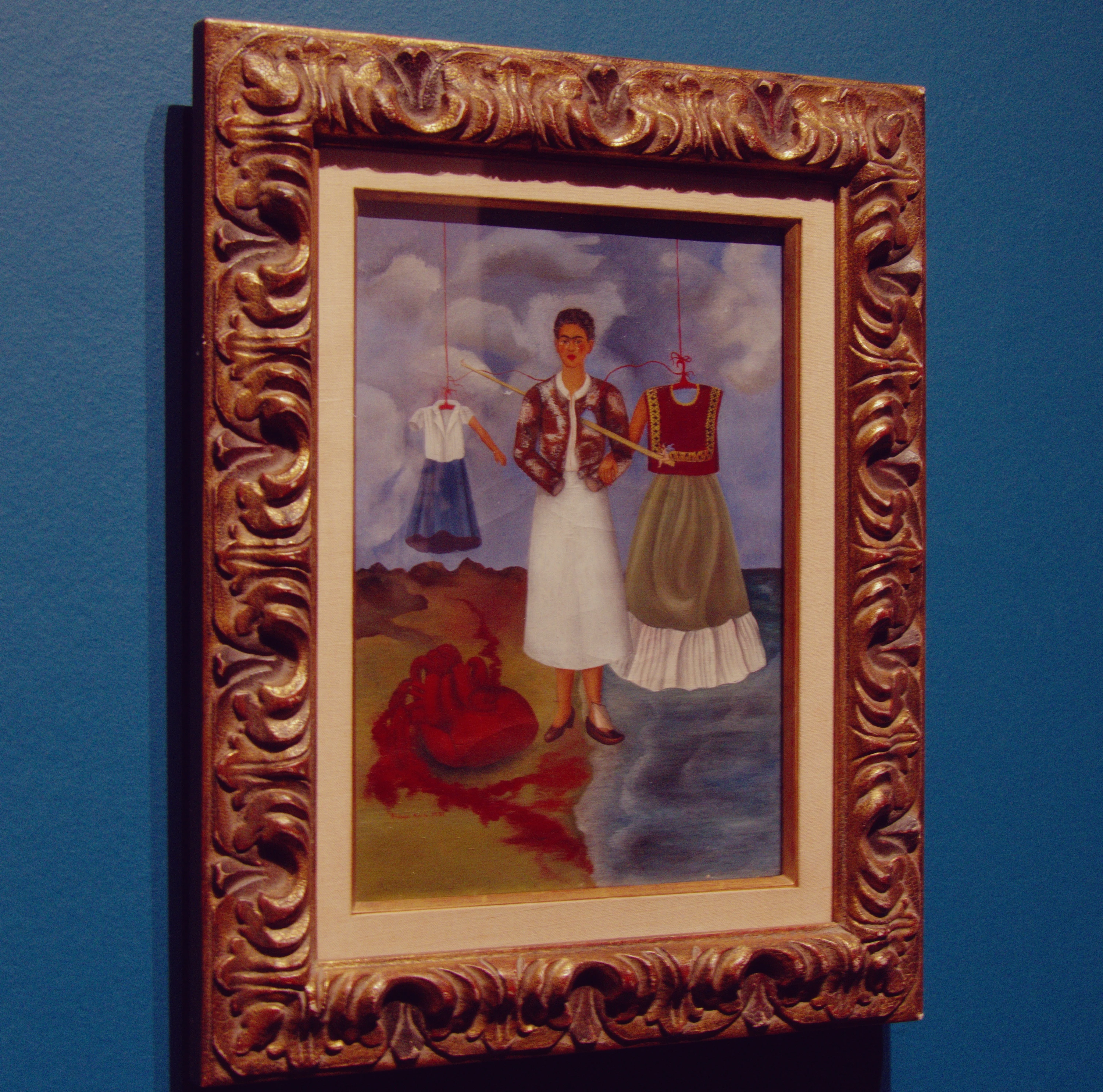 Memory, the Heart — Frida standing between a hanging schoolgirl dress and a Tehuana costume, a large heart bleeding on the ground